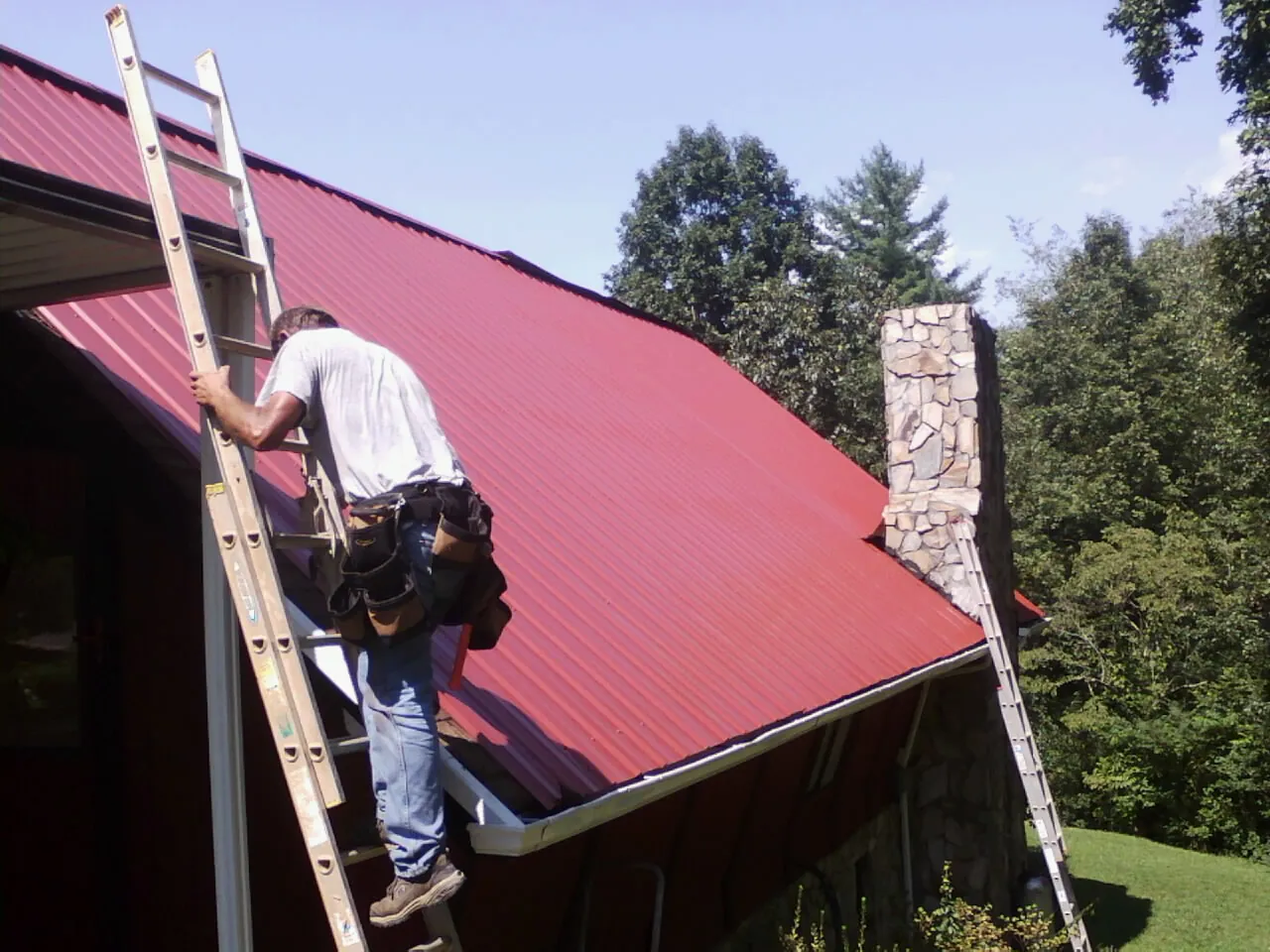Roofer on a ladder beside a red metal roof and stone chimney — Soffit & Fascia Repair by Roofing Brothers in El Sobrante